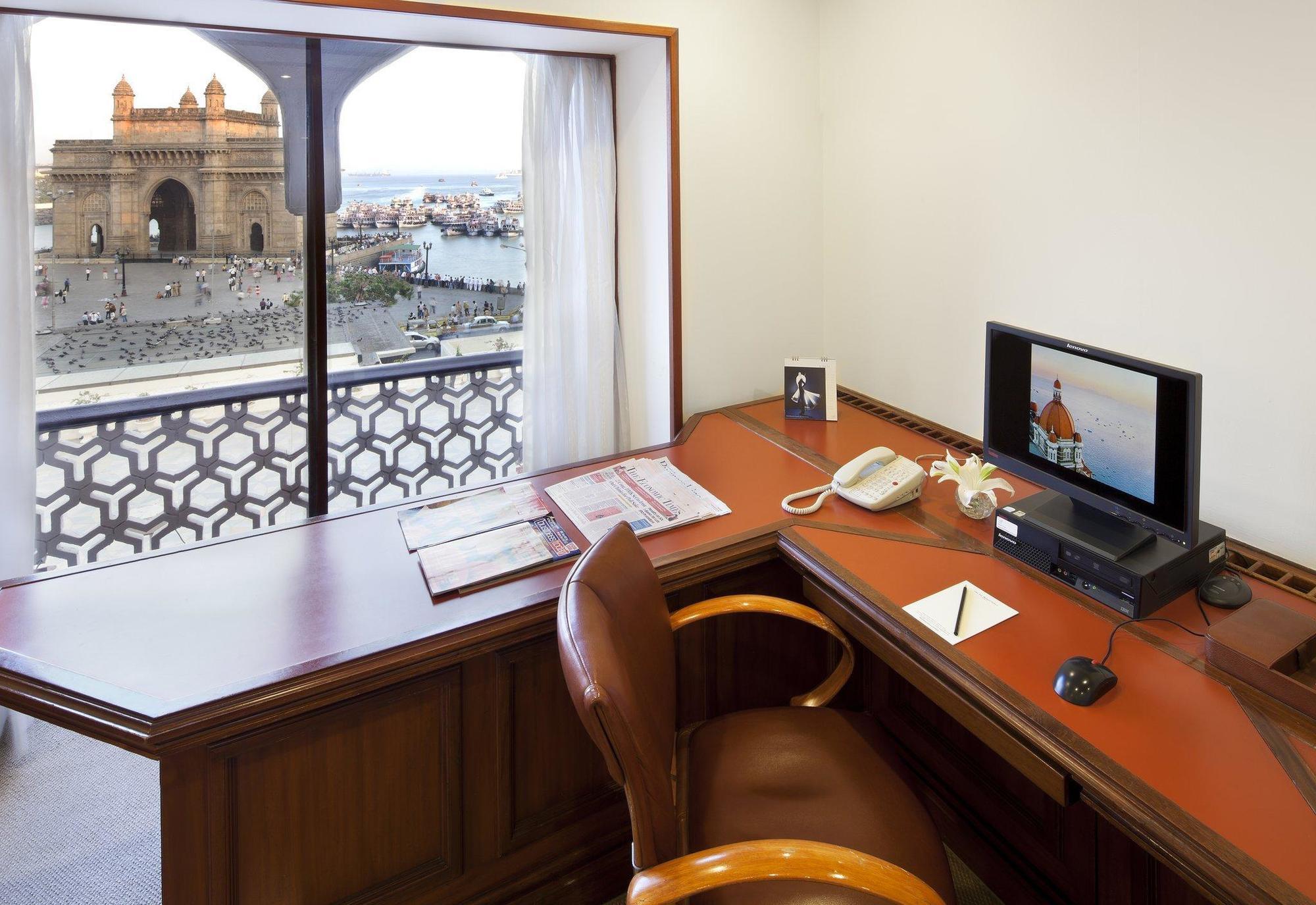 Hotel The Taj Mahal Tower, Bombay Esterno foto The photo shows a workspace with a wooden desk and an ergonomic chair. On the desk, there is a computer monitor, a telephone, and some stationery items. A window provides a view of an outdoor scene, possibly featuring water and structures or building