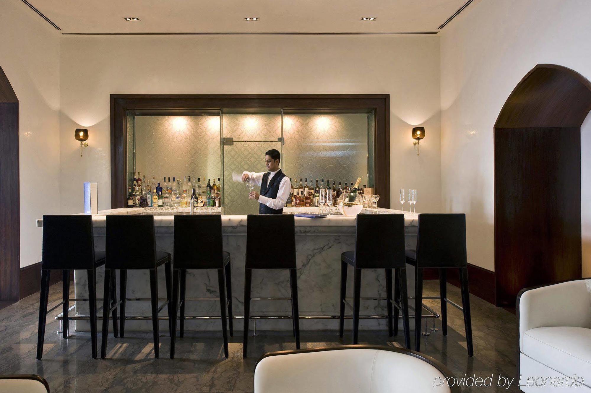 Hotel The Taj Mahal Tower, Bombay Ristorante foto The photo shows a stylish bar area. In the center, a bartender, dressed in a formal outfit, is standing behind a sleek, modern bar counter made of marble. The bar is lined with high chairs for patrons. Behind the bartender, there are shelves filled w